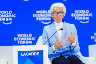 Christine Lagarde seated in front of a microphone and podium bearing her surname. Behind her is a blue background with 'World Economic Forum' written in white.