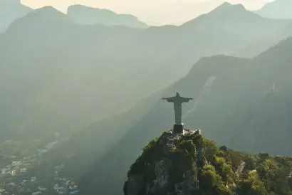The statue Christ the Redeemer sits on the summit of Sugarloaf Mountain. The peaks and slopes of other mountains in the background are partially obscured by haze.