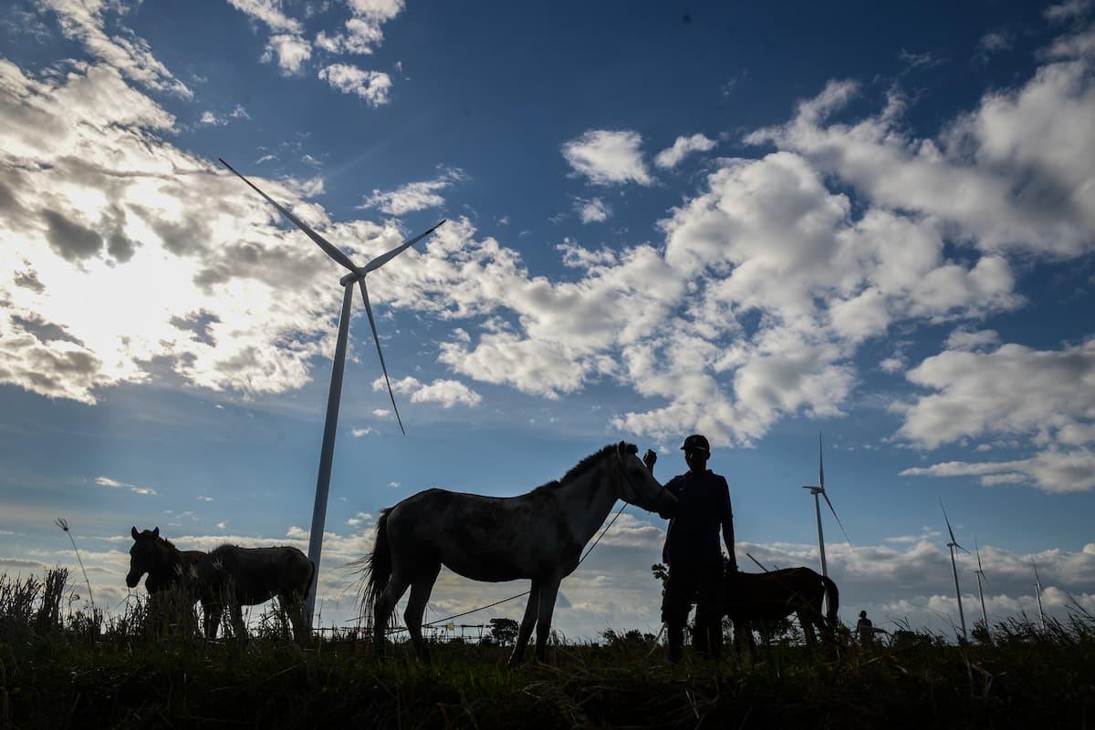 A person and four horse are silouhetted against a blue sky with smal clouds. Behoind them stand two wind power turbines.